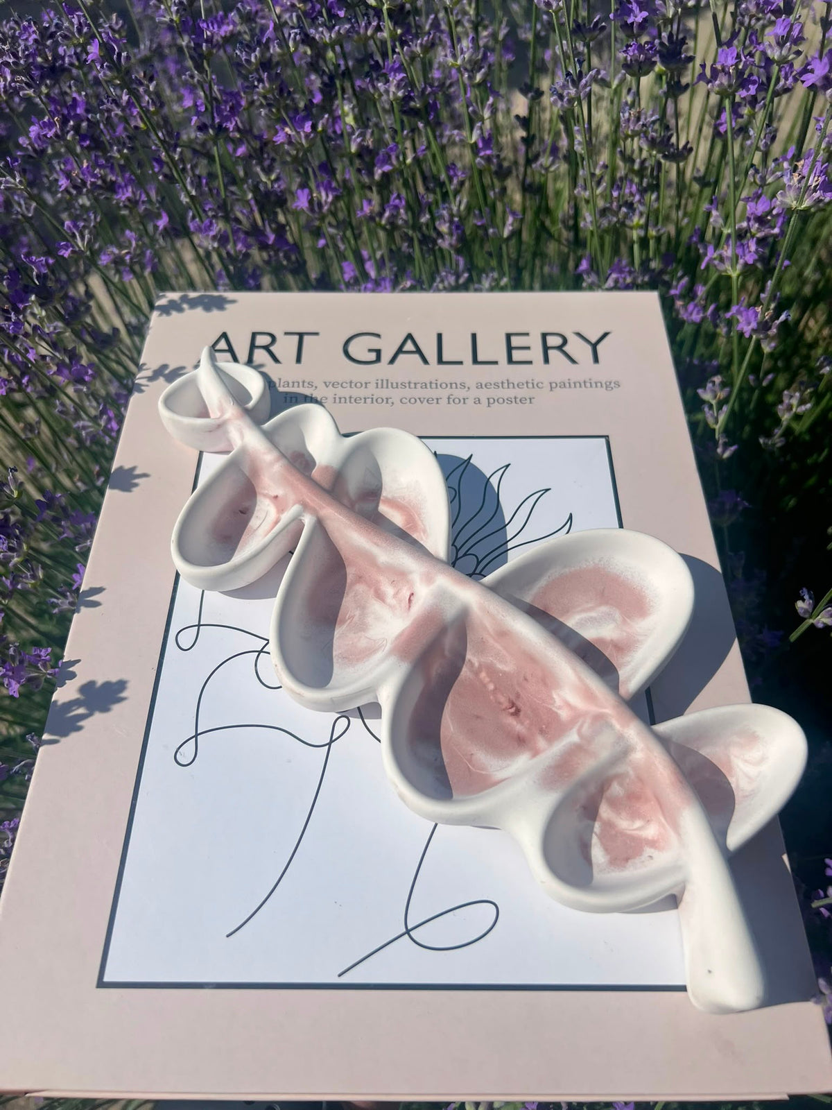 A white ceramic four-compartment dish with pink interior walls, placed on top of a printed poster in a real outdoor setting with lavender flowers in the background.